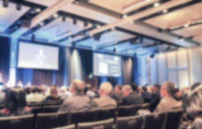 unscharfe Szene eines Seminarraums, in dem viele Menschen in mehreren Reihen sitzen und einem Redner zuhören.