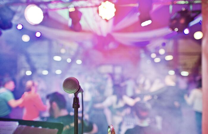 Event mit bunter, heller Beleuchtung, Menschen tanzen im Hintergrund, im Vordergrund ist ein Mikrofon zu sehen